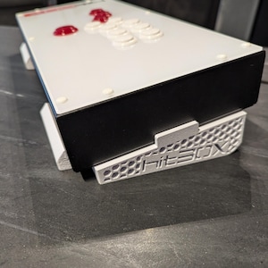 May include: A white arcade stick with red buttons and a black base. The stick is on a white stand with the word "Hitbox" printed on it.
