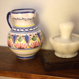May include: A decorative ceramic pitcher with a handle, featuring a floral design in pink, blue, yellow, and green. Beside it is a white marble mortar and pestle set, and a small wooden box.