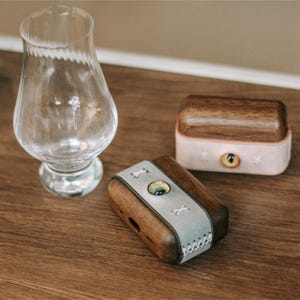 May include: Two small wooden boxes with leather accents. The boxes are closed and have a light brown wood finish. One box has a light grey leather accent with an X stitched into it. The other box has a pink leather accent with a small eye design.