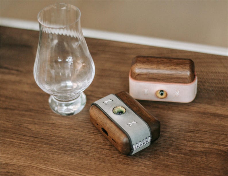 May include: Two small wooden boxes with leather accents. The boxes are closed and have a light brown wood finish. One box has a light grey leather accent with an X stitched into it. The other box has a pink leather accent with a small eye design.