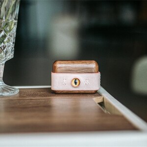May include: A small wooden box with a pink leather band and a gold eye design. The box is sitting on a wooden table.