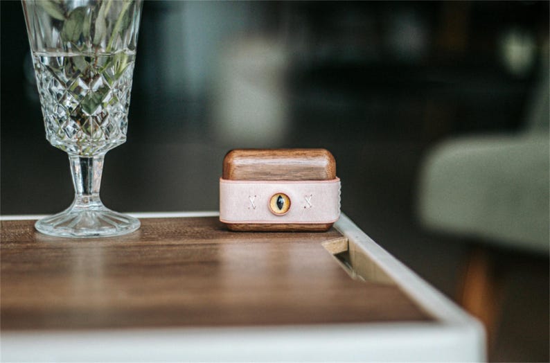 May include: A small wooden box with a pink leather band and a gold eye design. The box is sitting on a wooden table.