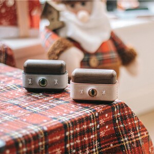 May include: Two small wooden boxes with lids, one is brown with a grey interior and the other is brown with a pink interior. Both boxes have a white 'X' on each side and a small round hole in the centre of the lid.