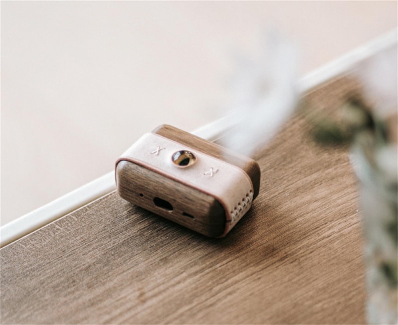 May include: A small, wooden and leather box with a light pink top and a round, silver button. The box has a small, rectangular opening on the side.
