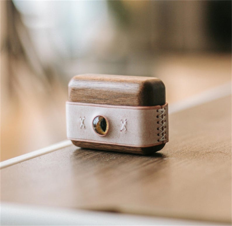 May include: A small wooden box with a light pink leather strap and a stitched design. The box has a round, gold-coloured eye in the centre of the strap.