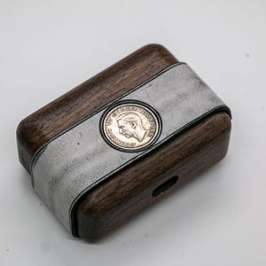 Puede incluir: Una pequeña caja de madera con una banda de cuero blanco y una moneda de plata incrustada en la parte superior. La moneda presenta un perfil de cabeza de hombre y el texto "W.D.G.R.  OMN. REX."