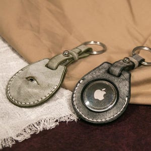 May include: Two leather keyrings with metal rings. One keyring is light green with a white stitch and the other is grey with a white stitch. The grey keyring has a clear plastic circle with a silver Apple logo inside.