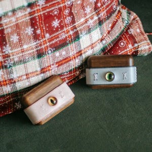 May include: Two small wooden boxes with leather lids. Each box has a single eye design on the lid. One box has a green eye and the other has a brown eye. The boxes are on a green surface with a red and white plaid fabric in the background.