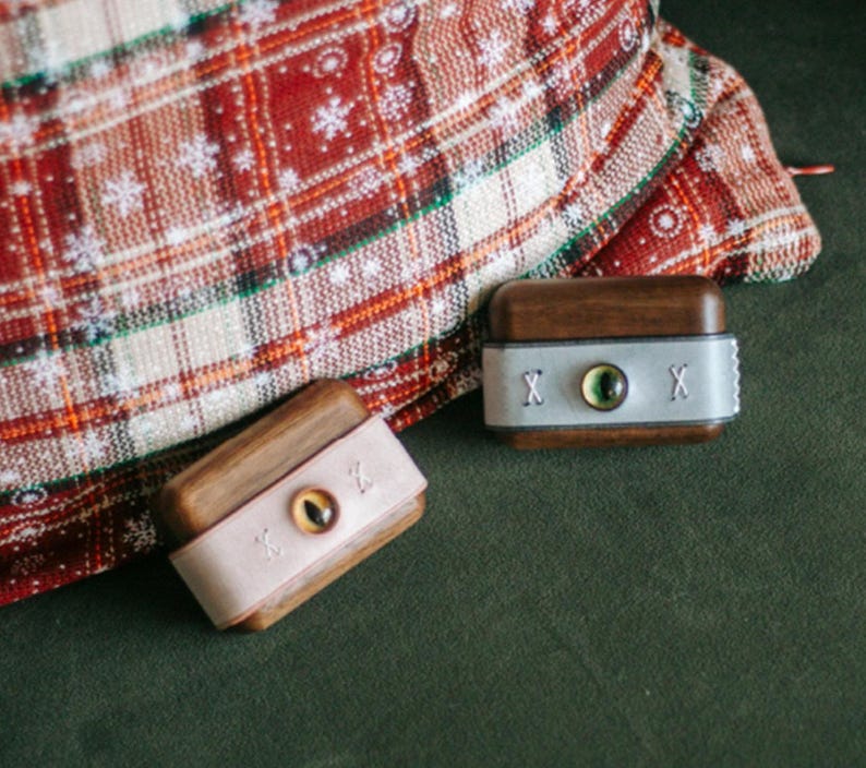 May include: Two small wooden boxes with leather lids. Each box has a single eye design on the lid. One box has a green eye and the other has a brown eye. The boxes are on a green surface with a red and white plaid fabric in the background.