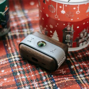 May include: A wooden and leather case for wireless earbuds with a green eye design and the letter "X" on each side.