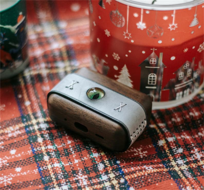 May include: A wooden and leather case for wireless earbuds with a green eye design and the letter "X" on each side.