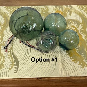 May include: Four spherical glass floats in varying shades of translucent green. One smaller float is encased in a dark gray netting. Attached to the glass floats are thin, multi-colored red, white, and black strings. The text Option #1 is visible.