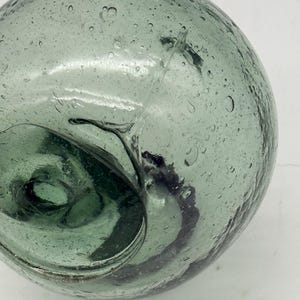 May include: A clear, round glass float with a seafoam green tint. The float has a spiral design on the inside and is covered in small bubbles. This decorative piece is ideal for nautical-themed decor.