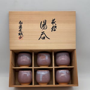 May include: A set of six small, round, purple-glazed ceramic tea cups in a wooden box. The box has Japanese characters printed on the lid. The cups are arranged in two rows of three, each with a slightly different glaze pattern.