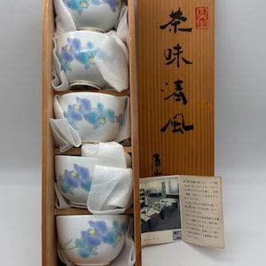 May include: A set of four white ceramic teacups with blue and purple floral designs, nestled in a wooden box. The box has a wooden lid with Japanese characters. The teacups are cushioned with white padding.