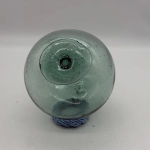 May include: A clear, round glass fishing float with a slightly green tint. The float has a small hole at the top and is attached to a blue and white rope base. This vintage nautical item is a decorative piece.