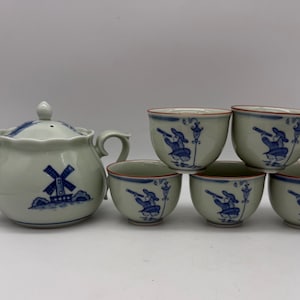 May include: A ceramic tea set featuring a teapot and four matching cups with a light gray glaze. Each piece is decorated with blue illustrations of a windmill and a figure holding a telescope, finished with a thin red rim around the edges.