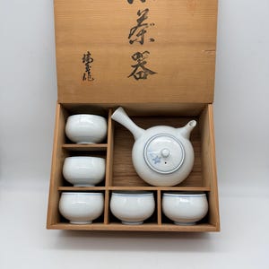 May include: A wooden box set containing a white ceramic teapot with a blue floral design and five matching teacups. The box has Japanese characters on the lid.