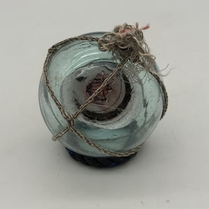Puede incluir: Un flotador de pesca de vidrio transparente con un tinte azul-verdoso, envuelto en una red de cuerda tejida. El flotador tiene un interior oscuro y está atado con una cuerda deshilachada en la parte superior. La base es de material tejido azul oscuro y negro.