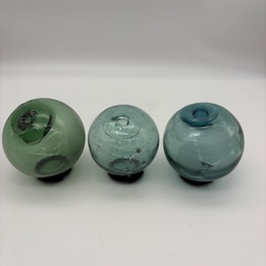 Puede incluir: Tres flotadores de pesca de vidrio en tonos verdes y azules. Cada flotador es redondo con una pequeña base oscura. El vidrio tiene un aspecto ligeramente texturizado, y los colores varían de un verde azulado intenso a un verde espuma de mar más claro.
