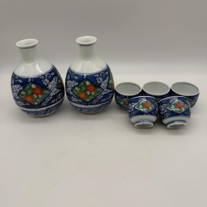 Puede incluir: Un juego de sake japonés con dos botellas de cerámica blancas y azules y seis tazas a juego. Las botellas y tazas tienen un diseño floral con detalles en rojo, verde y blanco. El juego está diseñado para servir sake.
