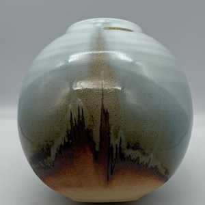 May include: A spherical ceramic vase with a smooth, glossy finish. The upper portion features a pale, light blue glaze that transitions into a darker, earthy brown tone at the base, characterized by vertical, drip-like streaks and speckles.