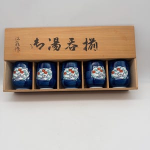 May include: A set of five blue and white ceramic tea cups with a floral design, presented in a wooden box. The cups feature a dark blue base with a white floral pattern and red accents. The wooden box has Japanese text.
