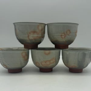 May include: A set of five ceramic tea bowls featuring a light gray glaze with subtle orange and brown mottled accents. Each bowl has a rounded body, a wide rim, and a distinct reddish-brown unglazed base, showcasing a smooth, fired finish.