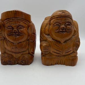 May include: Two wooden statues of smiling figures, carved from light brown wood. One figure holds a mallet, the other carries a large sack. The statues have detailed facial features and are likely decorative items.