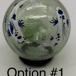 May include: A clear glass orb with a green tint, featuring hand-painted white koi fish with blue and black accents. The orb sits on a dark blue base. Text at the bottom reads "Option #1".