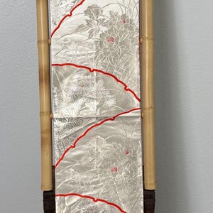 May include: A decorative textile with a floral pattern in shades of gray and white, accented with red outlines. The fabric is displayed vertically, supported by bamboo poles, and rests on a wooden chest.