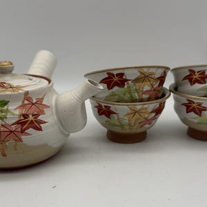 Puede incluir: Juego de té de cerámica con tetera y cuatro tazas a juego. El juego está decorado con un diseño de hojas de arce en rojo, verde y amarillo sobre un fondo blanco. La tetera tiene una tapa dorada y un asa curva.