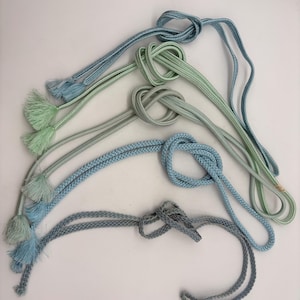 Puede incluir: Un surtido de cordones decorativos en tonos azules y verdes. Algunos cordones están anudados, mientras que otros tienen borlas en los extremos. Los cordones están dispuestos sobre una superficie blanca.