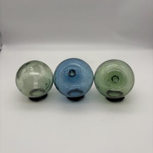 Puede incluir: Tres flotadores de pesca de vidrio en tonos verdes y azules. Cada esfera tiene un pequeño agujero en la parte superior y se asienta sobre una base oscura. Los flotadores están dispuestos en fila sobre un fondo blanco.