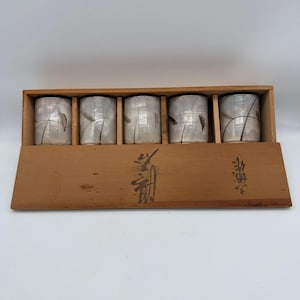 May include: A set of five ceramic cups with a light gray glaze and delicate black leaf designs, presented in a wooden box. The cups are cylindrical and arranged neatly within the box, which has Japanese characters printed on it.