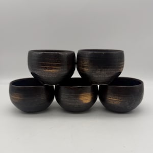 May include: A set of five small, round ceramic cups arranged in a pyramid formation. Each cup features a dark, matte black finish with horizontal bands of metallic gold or bronze glaze accents that wrap around the exterior surface of the vessels.