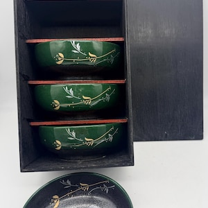 May include: A set of four green and black Japanese-style bowls with a floral design, presented in a black wooden box. The bowls have a red rim and are decorated with gold and white leaf patterns.