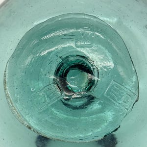 Könnte beinhalten: Nahaufnahme eines teal-farbenen Glasobjekts. Das Objekt hat eine runde Form mit einer kreisförmigen Vertiefung in der Mitte. Das Glas hat eine leicht strukturierte Oberfläche und ist transparent. Die mittlere Vertiefung hat einen dunkleren Teal-Farbton.