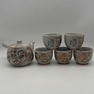 May include: A Japanese tea set featuring a teapot and six matching cups. The teapot and cups are white with colorful rooster and floral designs. The cups have Japanese characters on them.