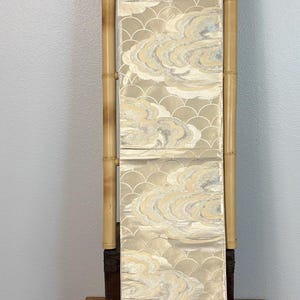 May include: A long, vertical textile panel featuring a repeating pattern of stylized clouds and overlapping semicircular wave motifs in shades of cream, beige, and metallic silver, suspended between two vertical bamboo poles.