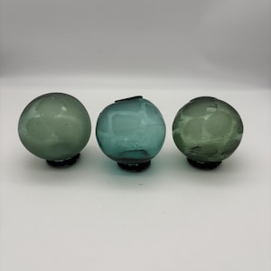 Puede incluir: Tres globos de vidrio en tonos verdes y turquesas. Cada globo tiene forma redondeada y se asienta sobre una base oscura. El vidrio tiene un aspecto ligeramente texturizado, y los globos están dispuestos en fila sobre un fondo blanco.