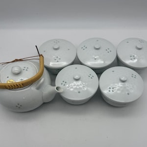 May include: A white ceramic tea set with a teapot and six matching cups. The teapot has a woven handle and a small spout. Each piece features a textured surface and a floral pattern. The set is designed for serving tea.