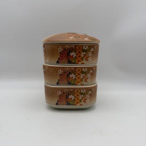 Puede incluir: Un recipiente cerámico apilable de tres niveles con tapa. El recipiente beige presenta un patrón floral y geométrico decorativo en tonos rojos, naranjas, verdes y blancos. La tapa está decorada con un diseño floral.