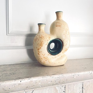 May include: A cream-coloured ceramic vase with a unique, organic shape. The vase features a large circular opening and two bottle-like forms connected together. It has a textured surface with brown accents and a teal-coloured interior.