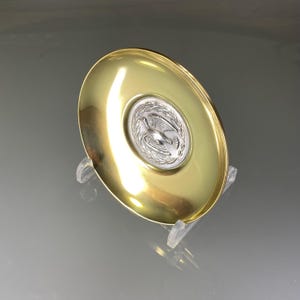 May include: A gold-colored metal dish with a silver-colored circular design in the center. The design features a stylized bird with wings spread, surrounded by a wreath.