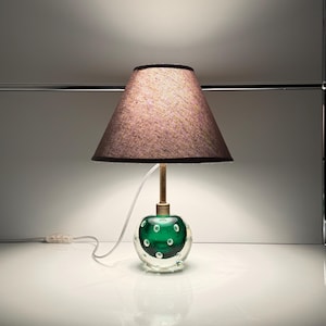 May include: A decorative table lamp featuring a green glass base with embedded air bubbles. The lamp has a brass stem and a mauve lampshade with a dark brown trim. The lamp is plugged into a white electrical outlet.