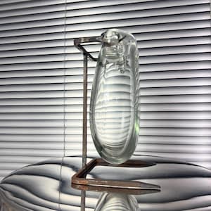 Può includere: Un vaso di vetro trasparente dalla forma allungata a goccia, tenuto da un supporto in metallo arrugginito. Il vaso ha una superficie liscia e riflettente con bolle interne. Il supporto ha una base quadrata e un supporto verticale.