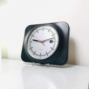 May include: A square, black-framed wall clock with a white face. The clock features black hour markers, black hands, and an orange second hand. The clock has a small date window at the 3 o'clock position and is labeled "se Quartz".