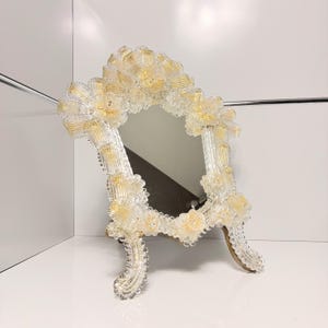 May include: A decorative vanity mirror with a clear glass frame adorned with gold and clear floral accents. The mirror has a rectangular shape with rounded edges and stands on two curved legs.