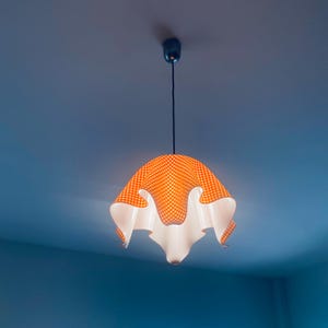 Puede incluir: Una lámpara colgante única, con una pantalla a cuadros naranja y blanca. La pantalla tiene un diseño ondulado y con volantes con un interior blanco. Cuelga de un cable negro y está montada en un techo azul.